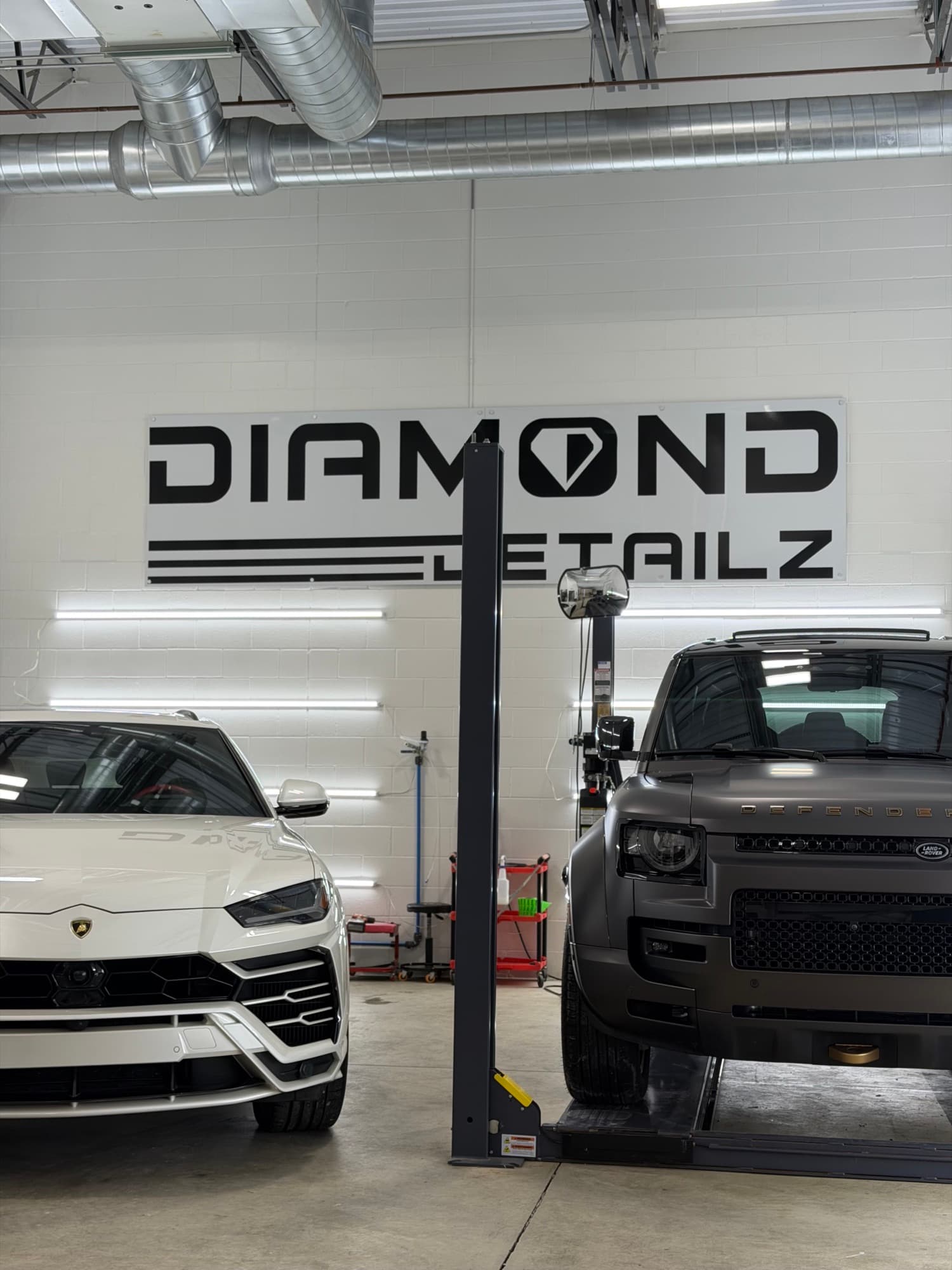 White Lamborghini Urus and matte-black Land Rover Defender side-by-side in the Diamond Detailz install bay