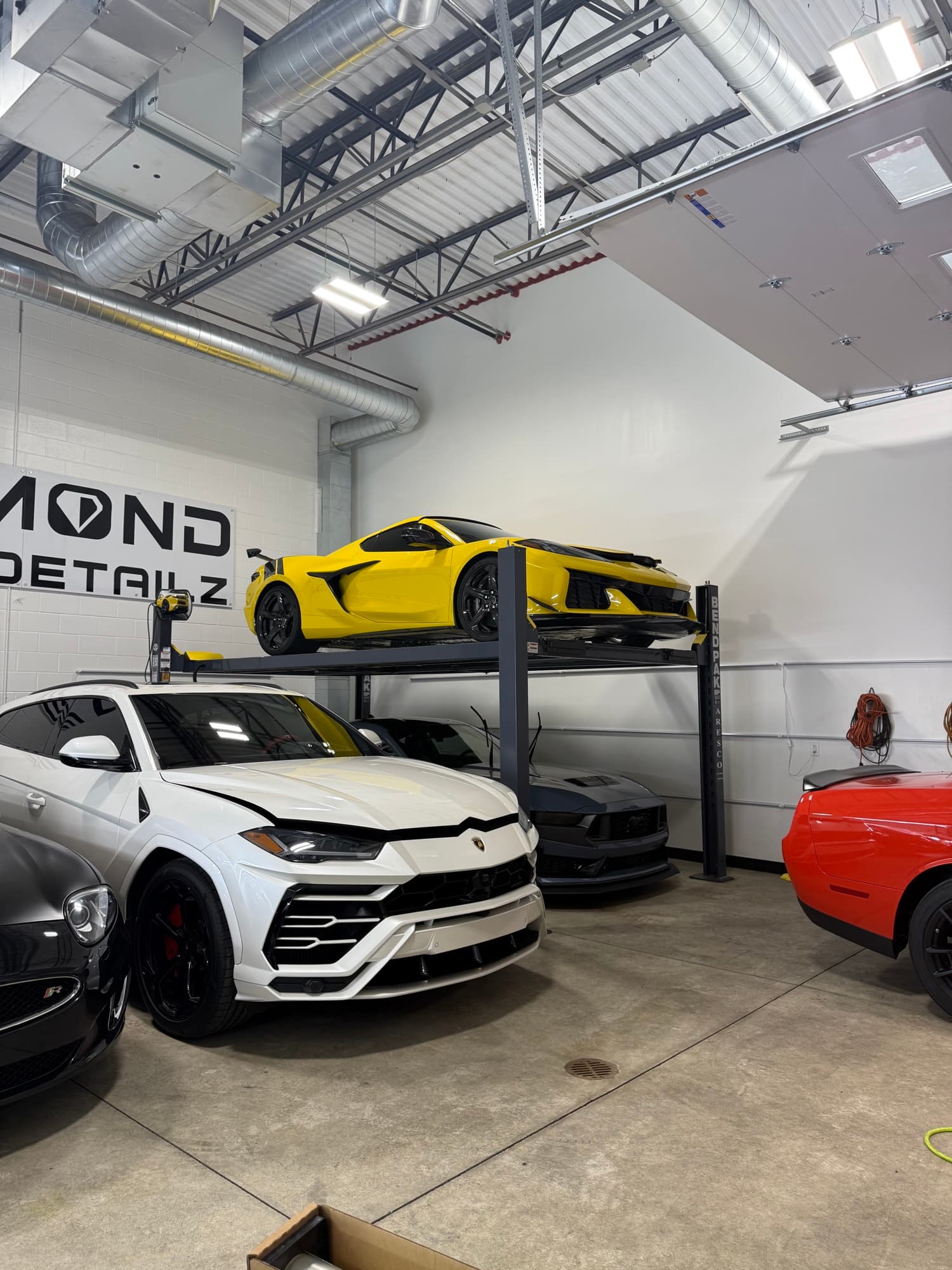 Yellow C8 Corvette on the lift above a white Lamborghini Urus inside the Diamond Detailz install bay